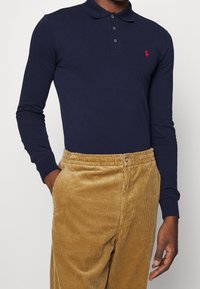 Camisa polo de manga larga azul marino con cuello y tres botones. Cuenta con un pequeño logotipo bordado en rojo en el pecho. Combinada con pantalones de pana color beige.