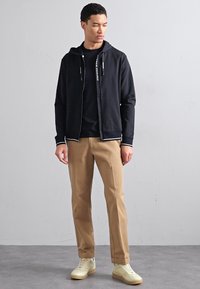 Felpa nera con zip e accenti bianchi, indossata sopra una t-shirt nera, abbinata a pantaloni beige e sneakers chiare, esposta su uno sfondo neutro.