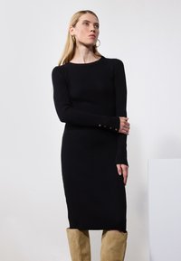 Svart ribbestrikket kjole med lange ermer, med knappdetaljer på ermene. Modellen er kombinert med lysebeige kne-høye støvler.