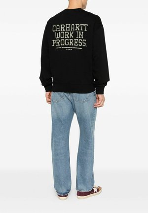 Persona che indossa una felpa nera con la scritta "CARHARTT WORK IN PROGRESS" sulla schiena, jeans azzurri chiari e scarpe da ginnastica marroni con una stella bianca.