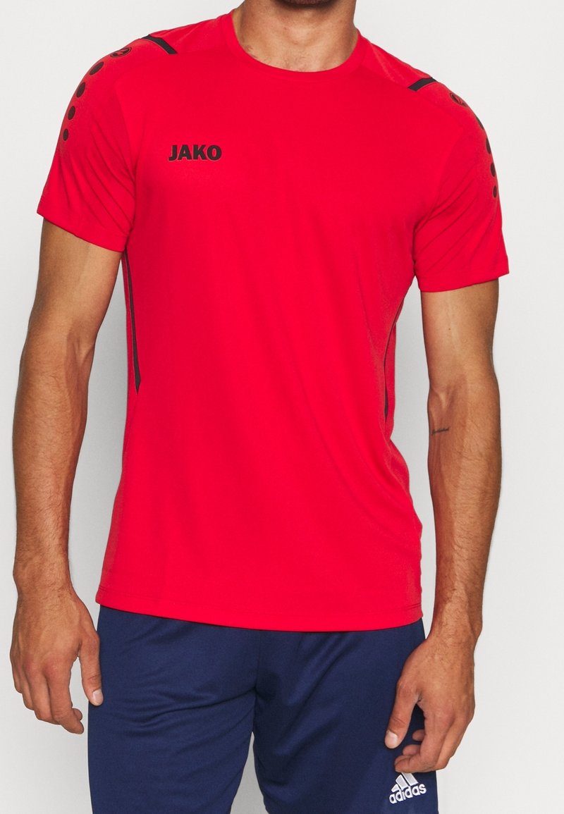 Hombre vestido con una camiseta deportiva JAKO de un brillante color rojo con detalles en negro en los hombros, combinada con pantalones cortos Adidas de color azul marino.