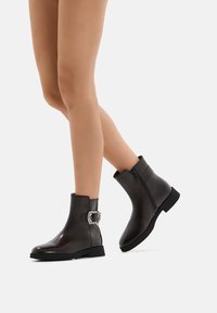 Stivaletti neri alla caviglia realizzati in materiale lucido, con cerniera laterale e un accento decorativo a fibbia. Suola piatta con un design a punta arrotondata.