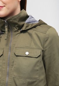 Olivově zelená bunda se zapínáním na zip, dvěma náprsními kapsami a šedou podšívkou kapuce. Texturovaná tkanina s viditelnými švy.