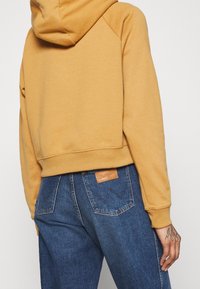 Zkrácená hořčicová mikina vyrobená z měkké látky s kapucí. V kombinaci s modrými džíny s vysokým pasem, které mají koženou značku na zadní straně.