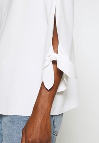 Hvid top med løstsiddende ærmer med slit og bundet stofdetaljer. Glat tekstur kombineret med lyseblå denim.