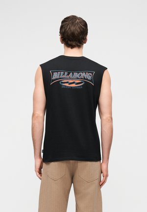 Vīrietis melnā bezroku krekla ar "Billabong" logotipu un tekstu "Since 1973 Australia" uz muguras, valkājot bēšas bikses.