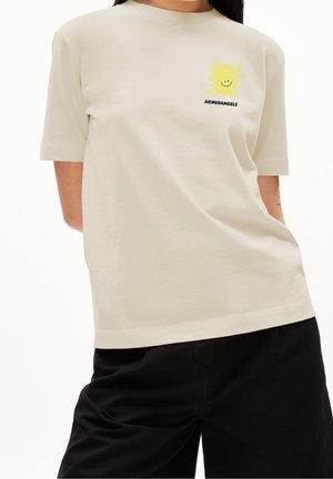 Personne portant un T-shirt beige avec un petit soleil jaune souriant et le texte "ARMEDANGELS" sur la poitrine, associé à un pantalon noir.