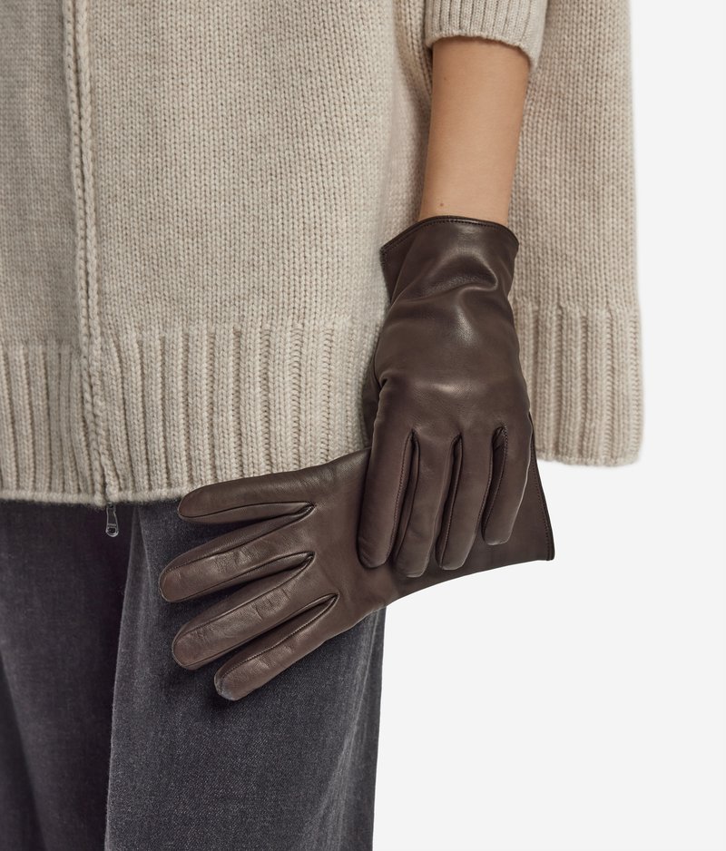 Personne portant un pull en maille beige et un pantalon foncé tient des gants en cuir marron foncé contre sa hanche.
