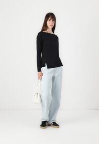 Black long-sleeve top with a boat neckline and side slits, paired with light blue wide-leg jeans and black sneakers. White handbag.