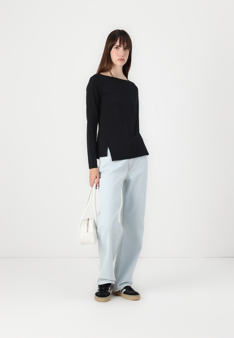 Schwarzes Langarmoberteil mit einem Boote-Ausschnitt und seitlichen Schlitzen, kombiniert mit hellblauen weitem Jeans und schwarzen Sneakern. Weißer Handtasche.