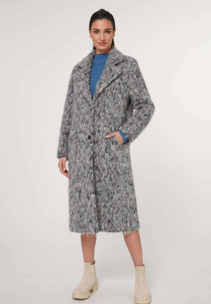 Grauer, strukturierter Mantel aus flauschigem Material mit langer Silhouette, Schalkragen und Knopfverschluss. Getragen über einem blauen Pullover und beigen Stiefeln.