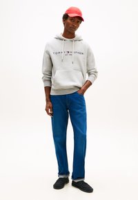 Sudadera con capucha gris con logo impreso, jeans azules de pierna recta y zapatos negros sin cordones. El modelo lleva una gorra roja. Fondo sencillo.