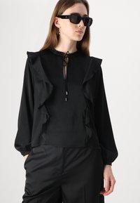 Blusa nera con dettagli a ruches, colletto alto e chiusura a fiocco. Maniche lunghe e un tessuto leggero e morbido. Indossata con pantaloni neri.