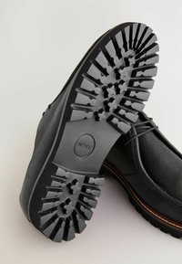 Chaussure noire en cuir avec tige texturée, bordure marron contrastante et semelle en caoutchouc robuste avec un motif en étoile pour une meilleure adhérence.