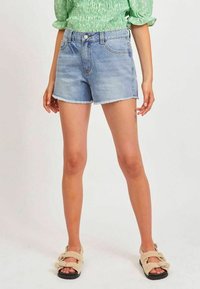 Jasnoniebieskie szorty dżinsowe z postrzępionym dołem, z klasycznym pięciokomorowym wzornictwem i zapięciem na guzik z przodu. Noszone z beżowymi sandałami.