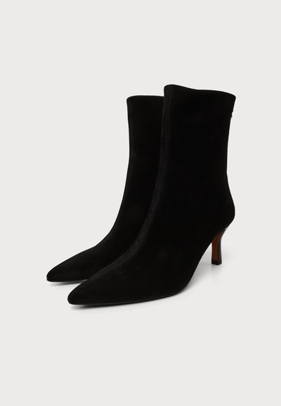 Paire de bottines en daim noir à bout pointu avec des talons fins de hauteur moyenne sur fond blanc.