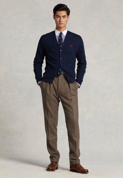 Cardigan in maglina blu navy con chiusura a bottoni, indossato sopra una camicia a righe e pantaloni a fantasia. Scarpe in pelle marrone completano l'abito.