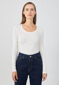 Maglietta bianca a maniche lunghe con scollatura a barca, abbinata a jeans a vita alta blu scuro con bottone frontale e cuciture in evidenza.