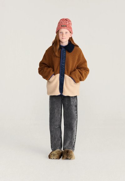 Enfant aux longs cheveux roux portant un bonnet rouge en tricot, une veste polaire marron et beige, un jean gris foncé et des chaussons marron duveteux, debout les mains dans les poches.
