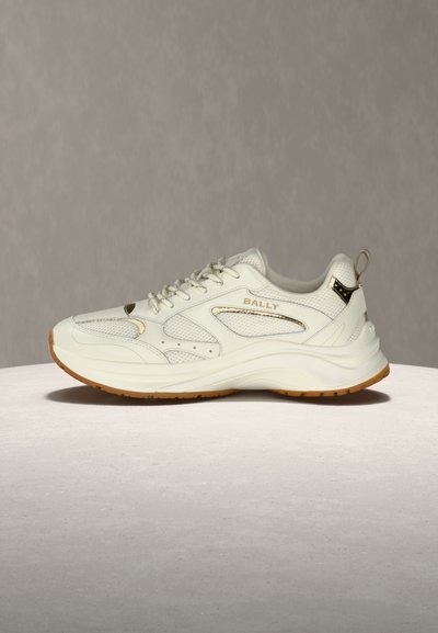 Λευκό αθλητικό παπούτσι Bally με πάνελ από δίχτυ και δέρμα, μπεζ λεπτομέρειες, παχιά σόλα και θηλιά, τοποθετημένο σε φωτεινή στρογγυλή επιφάνεια με φόντο γκρι.