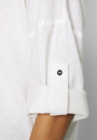 Camicia bianca di lino con maniche arrotolate, caratterizzata da un dettaglio in bottone nero sul polsino. Il tessuto ha un aspetto testurizzato e traspirante.