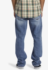 Jeans de mezclilla azul con un corte relajado, que presentan un desgaste sutil, bolsillos traseros cosidos y combinados con zapatillas negras y blancas.