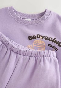 Lila souprava dětského oblečení s elastickým pasem a tričkem potištěným usmívajícím se pohárem na pití a textem "BABYCCINO TO GO."