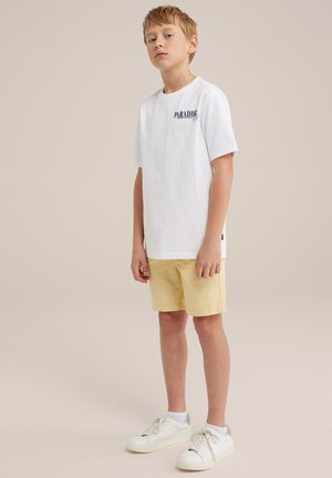 Camiseta de algodón blanca con "PARADISE" impreso en azul marino, combinada con pantalones cortos amarillos y zapatillas blancas. Diseño simple y casual con mangas cortas.