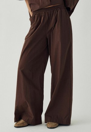 Brune viddebukser med elastisk taljebånd båret med tan moccasin-stil sko på lys baggrund.