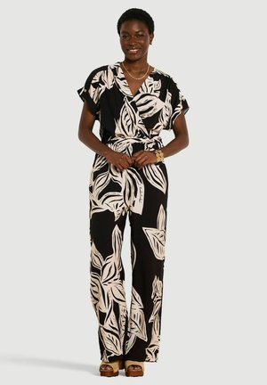 Femme souriante portant une combinaison imprimée de feuilles noire et blanche, à manches courtes, col en V, jambes larges, et sandales compensées beiges.