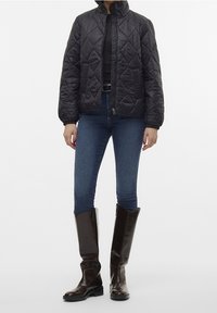 Veste matelassée noire avec un col montant, associée à un jean skinny bleu foncé et des bottes en cuir marron montant jusqu'au genou.