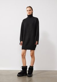 Zwarte oversized trui jurk met een hoge hals, geribde manchetten en zoom; gecombineerd met stevige zwarte laarzen met zachte stof en dikke zolen.