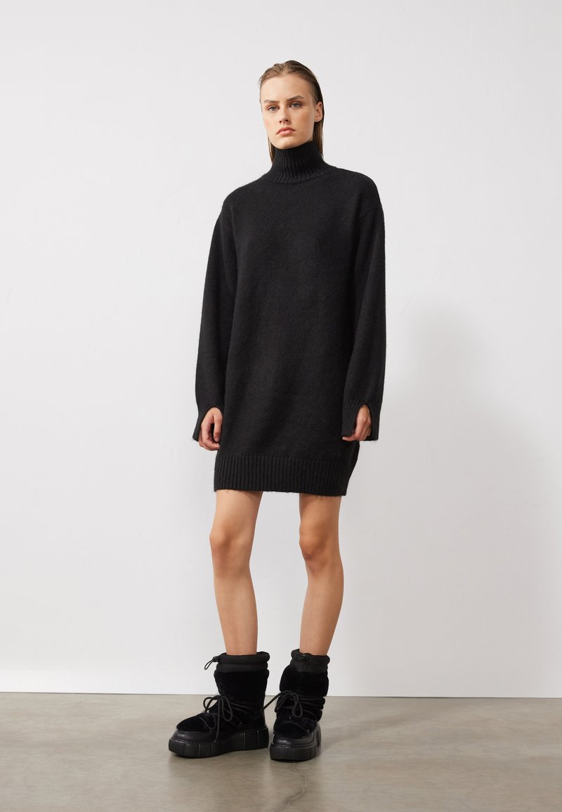 Zwarte oversized trui jurk met een hoge hals, geribde manchetten en zoom; gecombineerd met stevige zwarte laarzen met zachte stof en dikke zolen.