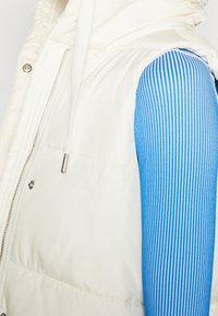 White puffer vest with a zipper, featuring a high collar and shiny silver hardware. A blue and white striped long-sleeve top is partially visible.