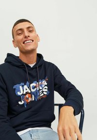 Sudadera marina con texto gráfico en blanco y naranja "JACK & JONES". Presenta capucha con cordón, puños acanalados y una textura suave. Modelo sentado.