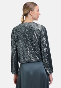 Schwarze Pailletten-Cropped-Bluse mit langen Ärmeln und rundem Ausschnitt. Glattes Material, das das Licht reflektiert, getragen über einem grauen Rock.