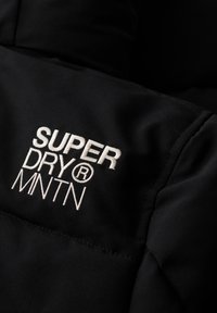 Zwarte pufferjack met witte geborduurde "SUPER DRY MNTN"-logo, zachte textuur en gestikte ontwerp voor isolatie en warmte.