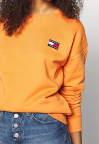 Oranžový svetr vyrobený z měkkého materiálu, který má malou značkovou nášivku Tommy Jeans v černé, červené a bílé barvě. Spárováno s modrými denimovými šortkami.