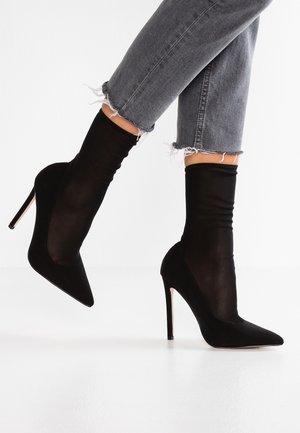 Nogi w przetartych szarych dżinsach i czarnych botkach typu sock boots na szpilce z zaostrzonym noskiem, jedna stopa uniesiona pokazująca kształt buta i wysokość obcasa.