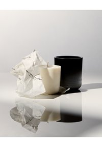 Schwarzer, zylindrischer Kerzenhalter neben einer weißen Wachskerze, umwickelt mit zerknittertem Papier mit gedrucktem Branding, das sich auf einer glänzenden Oberfläche spiegelt.