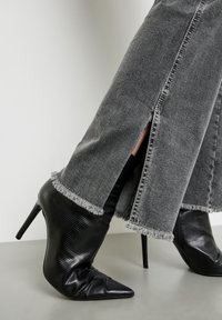 Botines negros de tacón con punta afilada y acabado texturizado, combinados con pantalones de mezclilla gris con dobladillos deshilachados y hendiduras laterales.