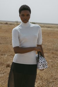 Šviesiai pilkas trumpomis rankovėmis megztinis su aukštu kaklu, derinamas su juoda permatoma sijonu ir balta rankine su juodais taškais.