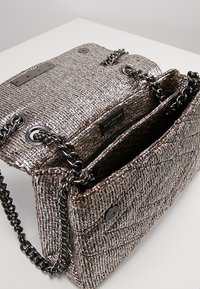 Bolso de mano metálico tejido con textura acolchada, color plata, con cadena, dos compartimentos y etiqueta de marca que presenta "Kurt Geiger."
