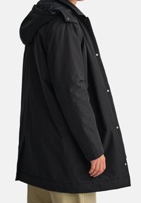 Sort vandtæt jakke med hætte, der har en quiltet foring, trykknapper og en lige skæring. Materialet ser glat og slidstærkt ud.