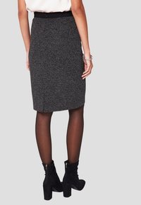 Mujer con una falda lápiz texturizada de color gris oscuro a la altura de la rodilla, pantys negros transparentes y botines negros de tacón, de pie frente a un fondo liso.