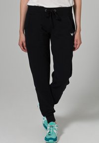 Pantalones jogger negros con una cinturilla elástica, cordón, bolsillos laterales y un pequeño detalle de logo. Combinados con zapatillas deportivas en color teal.