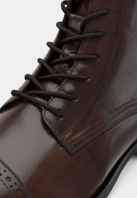 Καφέ δερμάτινη μποτάκια με γυαλιστερή φινέτσα, σχέδιο με κορδόνια και λεπτομερή ραφή κατά μήκος των πλαϊνών και της μύτης.