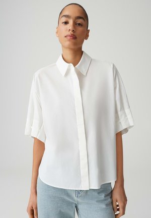 Jeune personne aux cheveux très courts portant une chemise blanche ample à manches courtes avec boutons et un jean bleu clair, debout devant un fond gris.