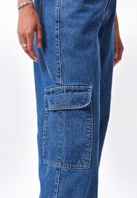 Pantalones cargo de mezclilla en azul medio con un corte holgado. Cuenta con un bolsillo lateral con tapa y costuras visibles para mayor durabilidad.