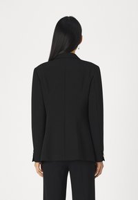 Veste noire ajustée avec une silhouette structurée, des revers crantés et des détails de couture subtils. Tissu lisse avec une finition mate.